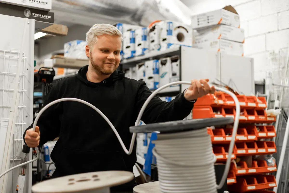 En mann med lyst hår håndterer en kabelrull inni et lagerrom fylt med utstyr og hyller.