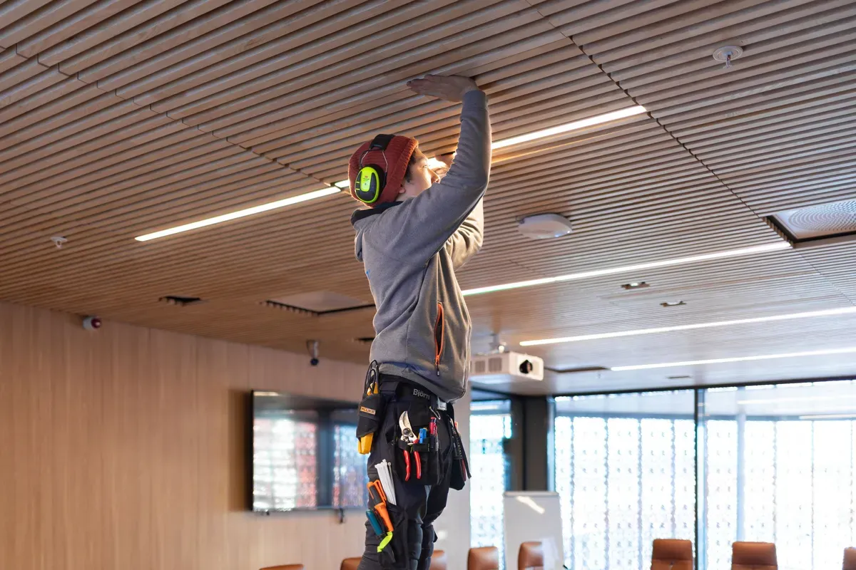 En håndverker med verktøybelte og hjelm jobber i taket på et moderne møterom.