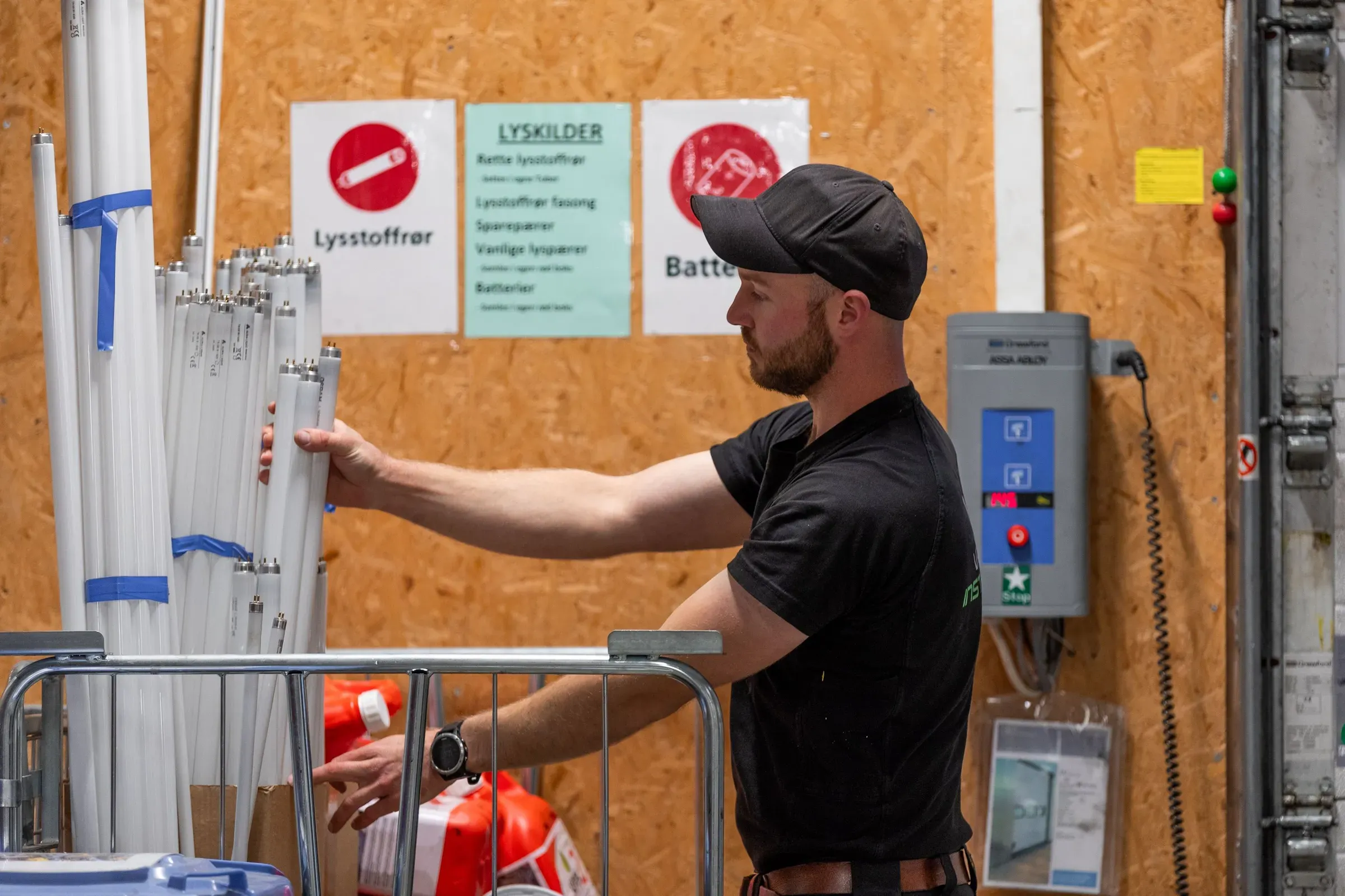 En mann med svart caps og sort t-skjorte sorterer brukte lysrør ved en gjenvinningsstasjon. Bak ham er det treplater og instruksjonsskilt om resirkulering av lyspærer og batterier.
