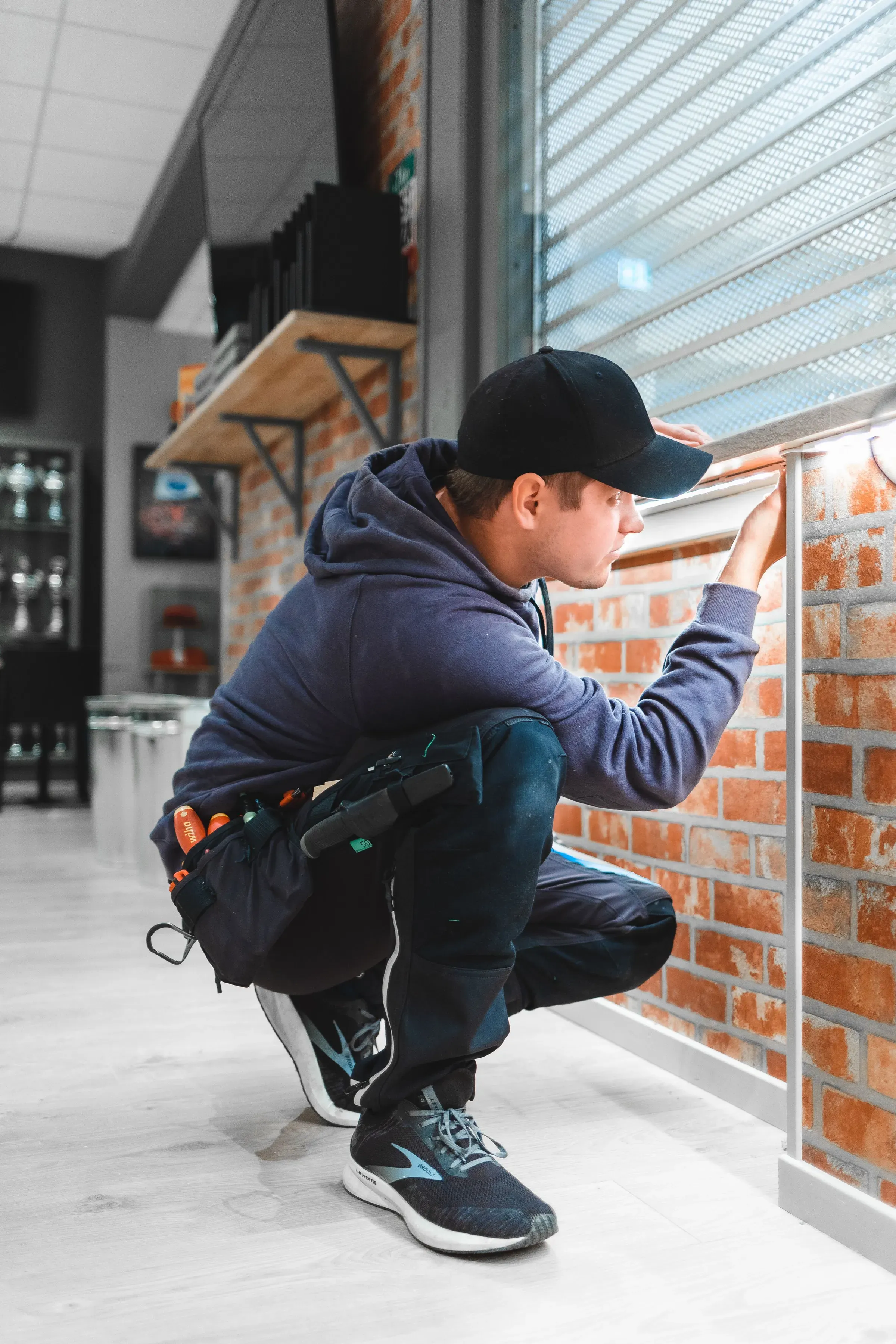 En håndverker i blå hettegenser og svart caps jobber ved et vindu inne i et rom med mursteinsvegg. Han har verktøy i beltet og undersøker vindusrammen mens han sitter på huk.