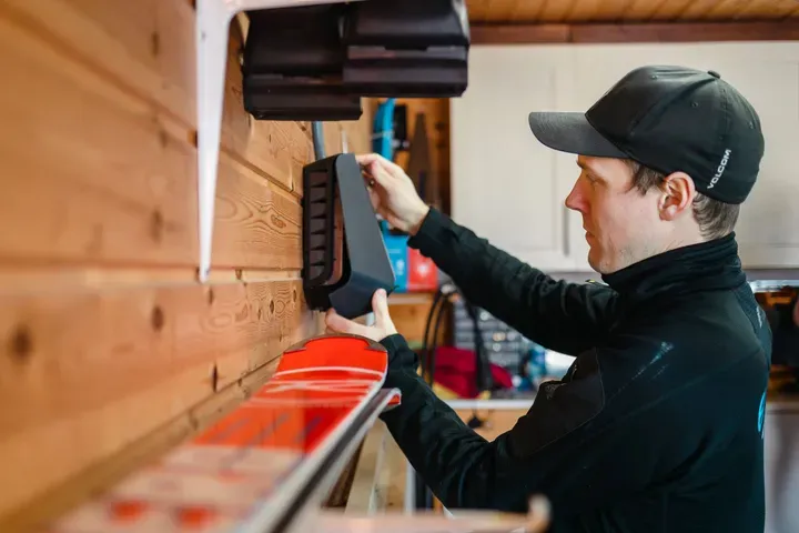 En mann i svart jakke og caps plasserer en svart boks på en hylle i et rom med trebvegger. Foran ham står et par røde og hvite ski.
