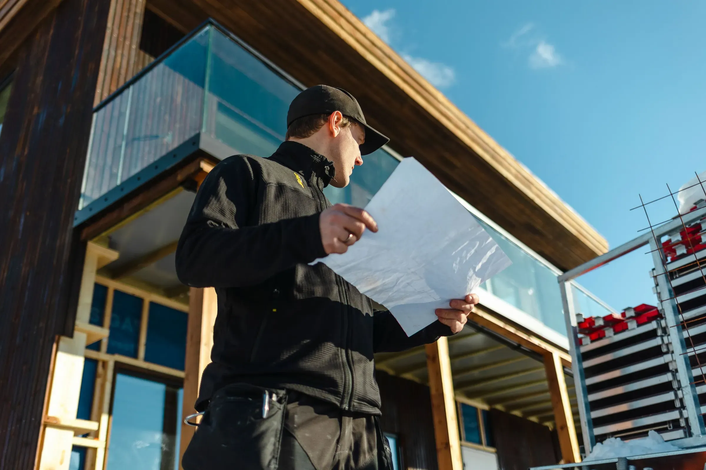 En mann i arbeidsklær holder byggeplaner foran et moderne hus under konstruksjon.