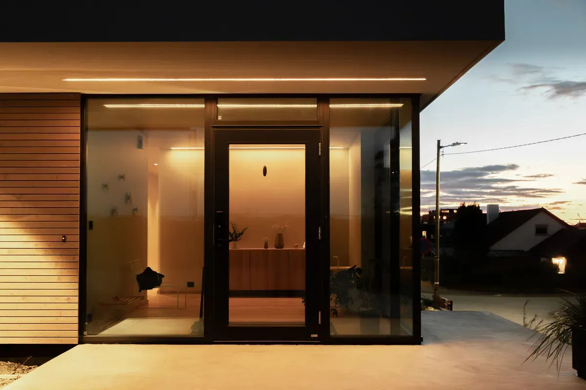 Moderne inngangsdør med store glassvinduer på begge sider av døren, huset er godt opplyst innvendig mens det er skumring ute.