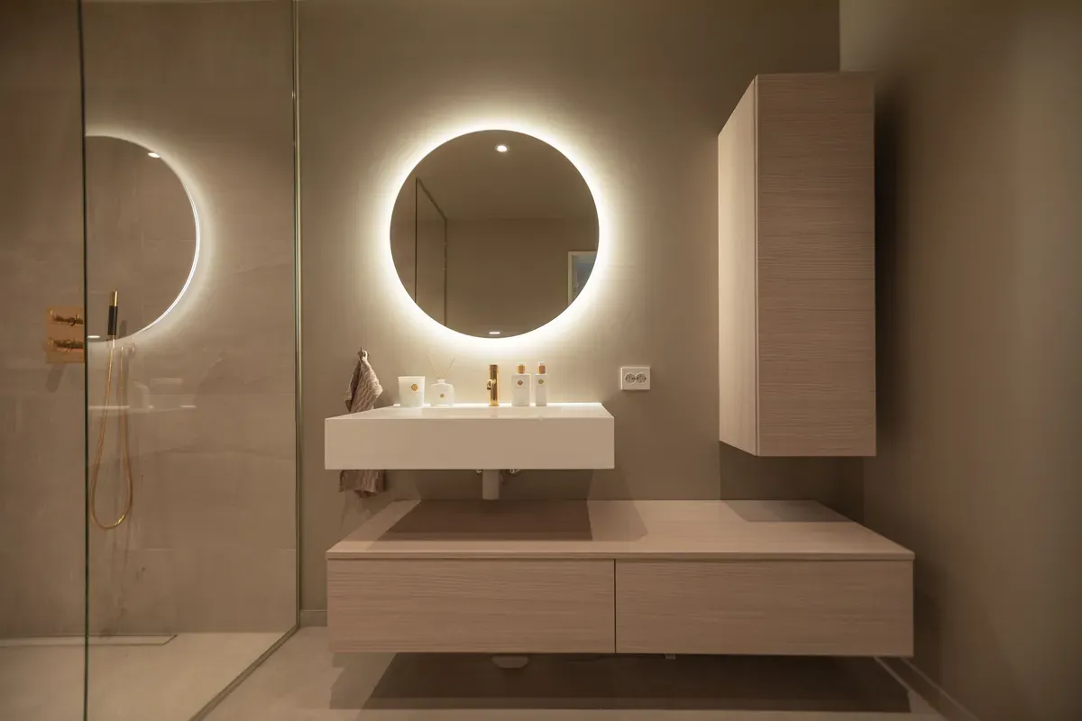 Moderne baderom med runde speil med bakgrunnsbelysning, vegghengt servantskap og dusjhjørne til venstre.