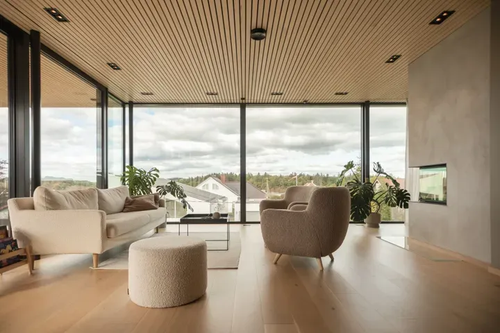 Lyst og moderne stue med store vinduer, lyse tremøbler og utsikt mot naturen utenfor.