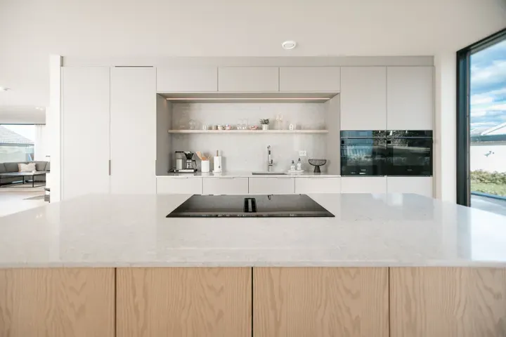 Moderne og minimalistisk kjøkken med lys benkeplate, innebygd koketopp og åpne hyller med kjøkkenutstyr.