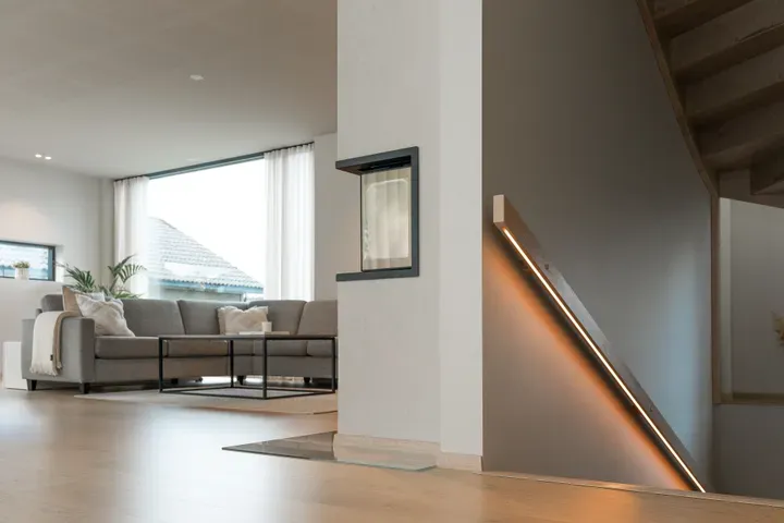 Moderne stue med lyse tregulv, stor sofa, glassbord og trapp med integrert LED-belysning.