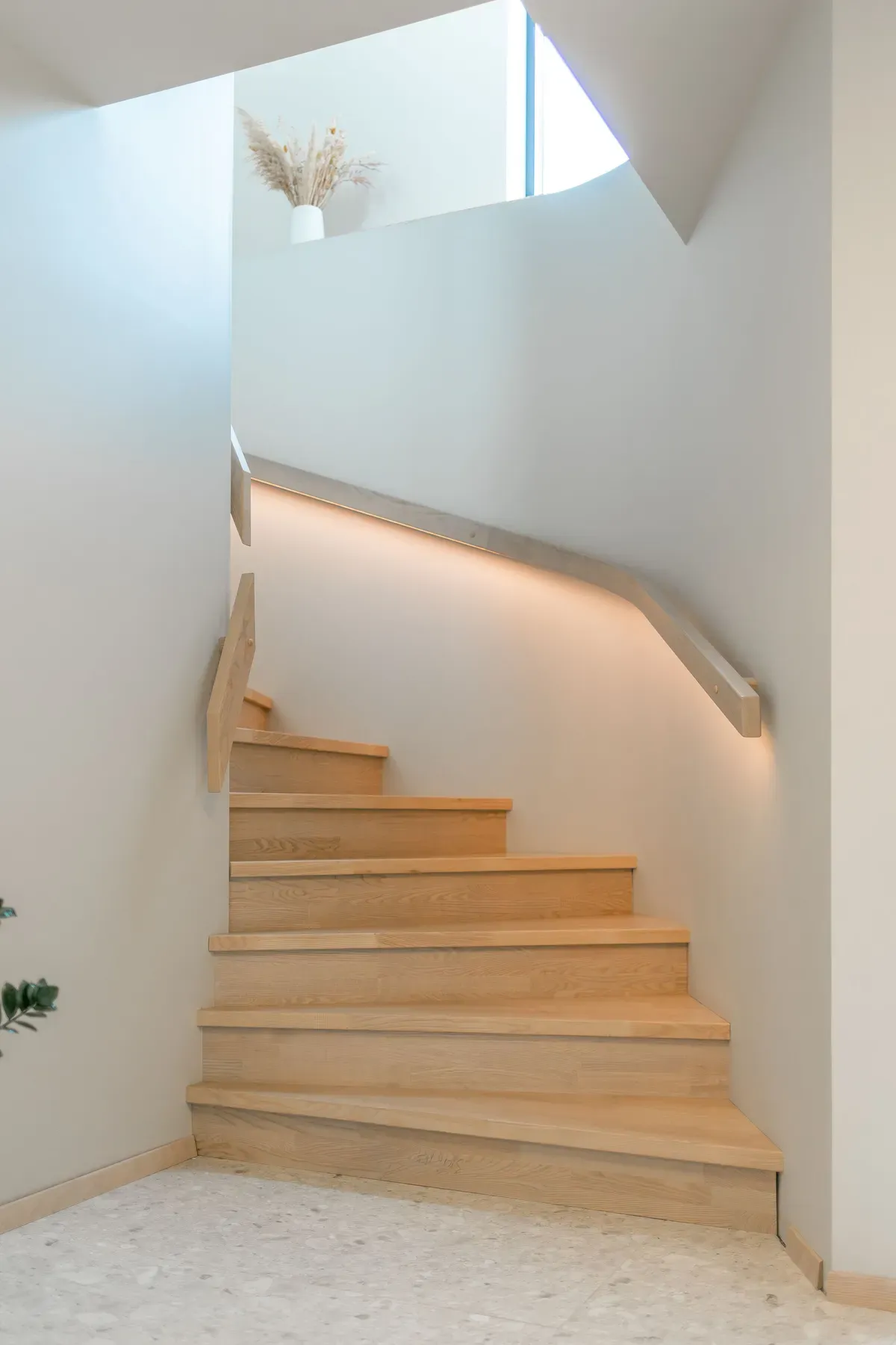 En moderne tretrapp med lyse tretrinn som svinger mot venstre, hvite vegger, en enkel håndlist og indirekte belysning under rekverket. Øverst i bildet står en hvit vase med tørkede strå foran et vindu.