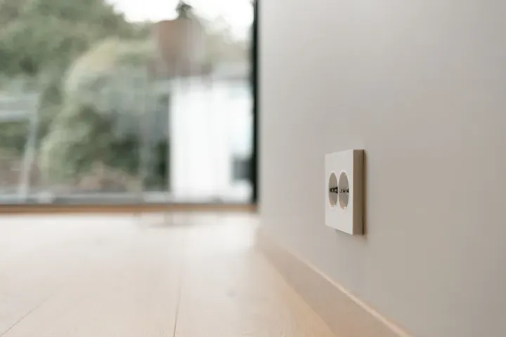 Nærbilde av en dobbel stikkontakt på en lys vegg i et moderne rom med tregulv. Utsikt til hage eller uteområde gjennom et stort vindu i bakgrunnen.