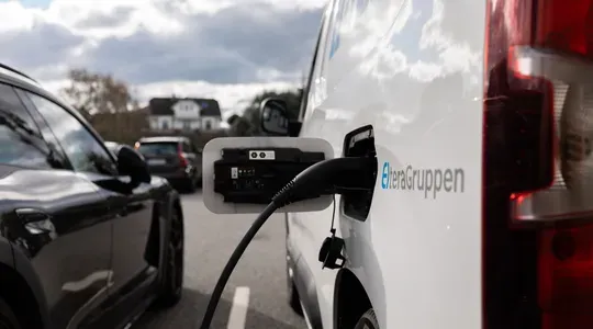 En hvit varebil lades med en ladekabel koblet til strømuttaket. Nærbilde av bilen med ladestasjon i fokus, og andre biler parkerte i bakgrunnen.
