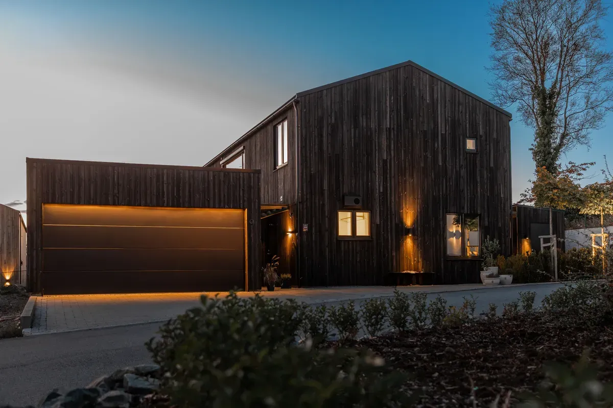 Et moderne, mørkt trekledd hus med innfelte utelys og tilhørende garasje, avbildet i skumringen.