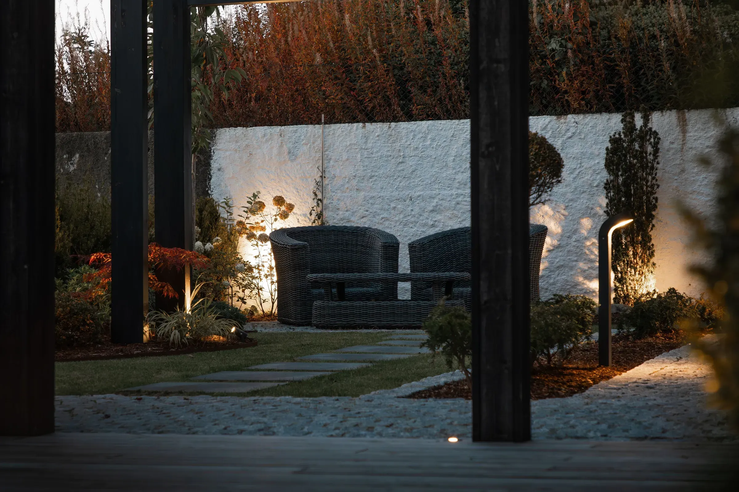 To mørke rottingstoler står mot en hvit murvegg i en hage, omgitt av planter og myk belysning om kvelden.