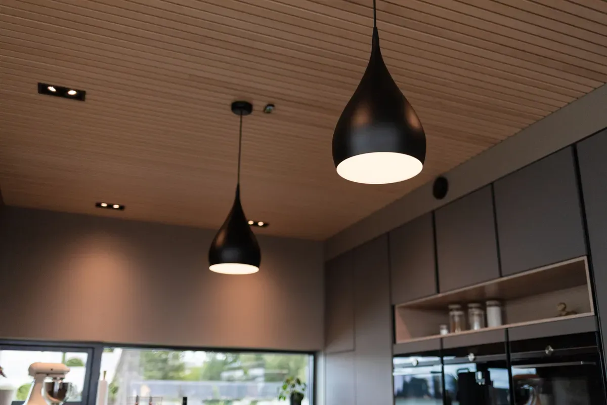 To moderne, hengende taklamper i svart over et kjøkken med grå skap og paneltak.