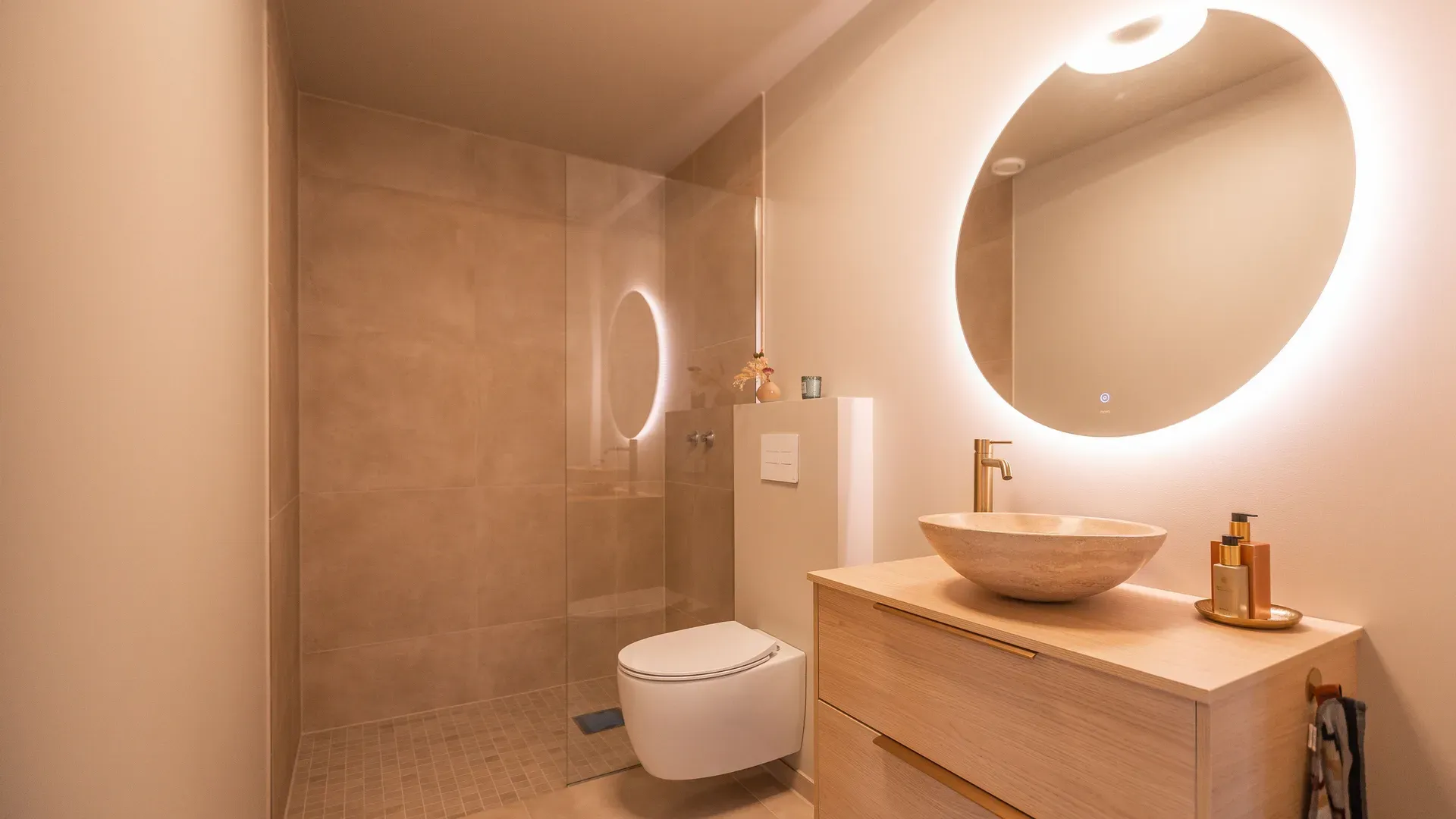 Et moderne bad med dusj, vegghengt toalett og servant på et lyst tremøbel. Det runde speilet har innebygd LED-belysning.