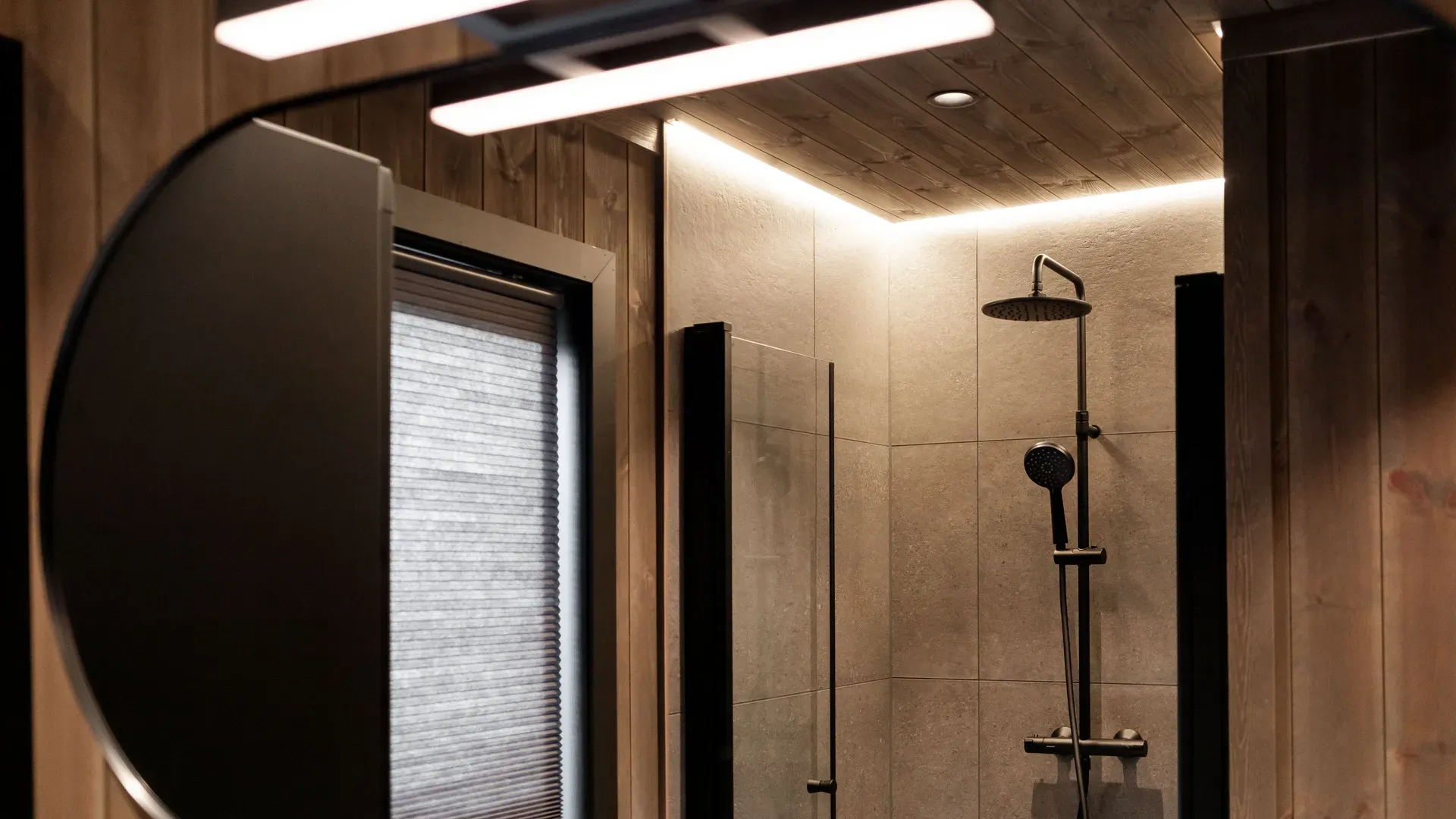 Moderne baderom med dusjhjørne, synlig gjennom et speil. Trehvit vegg og tak, belysning over speilet og dusjhode synlig i bakgrunnen.