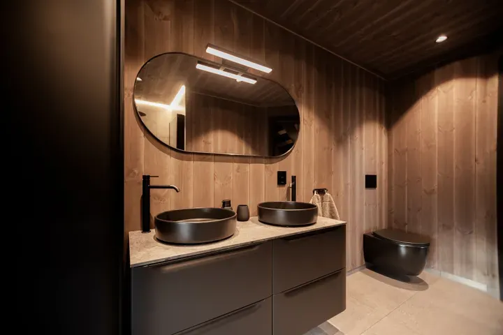 Moderne baderom med to svarte servanter, ovalt speil, mørke armaturer og veggmontert toalett. Veggene er i naturlig trepanel.