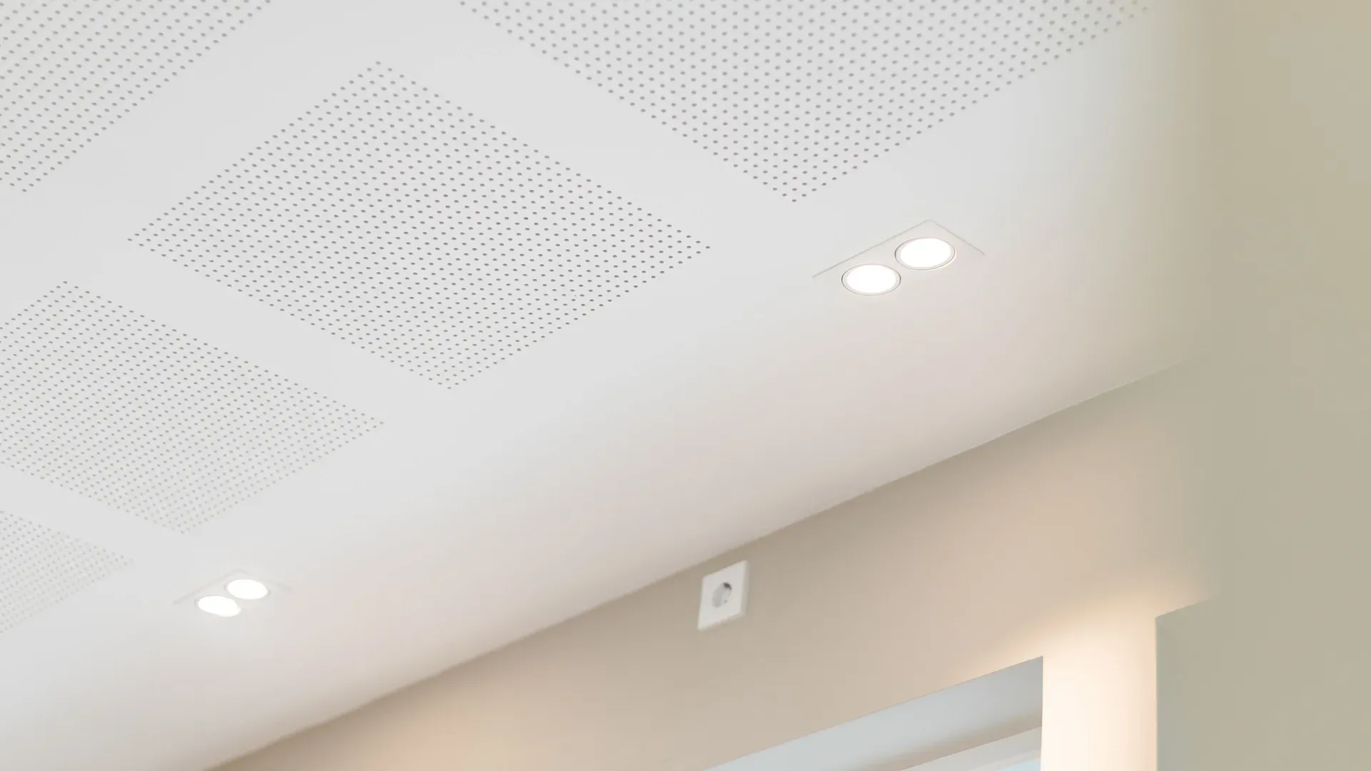 Et lyst tak med integrerte spotlys og perforerte lydplater. Under taket ses en vegg med et vindu og vegguttak.