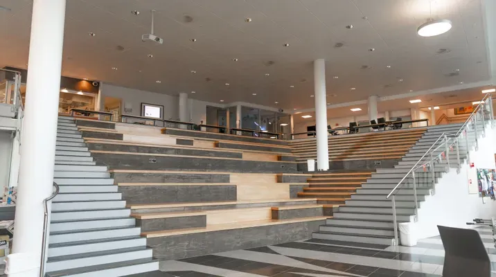 Et moderne innendørs amfiområde med brede trappeavsats og sitteplasser, omgitt av glassrekkverk og lyse søyler. Tepoer sitter gjerne folk til møter eller foredrag.