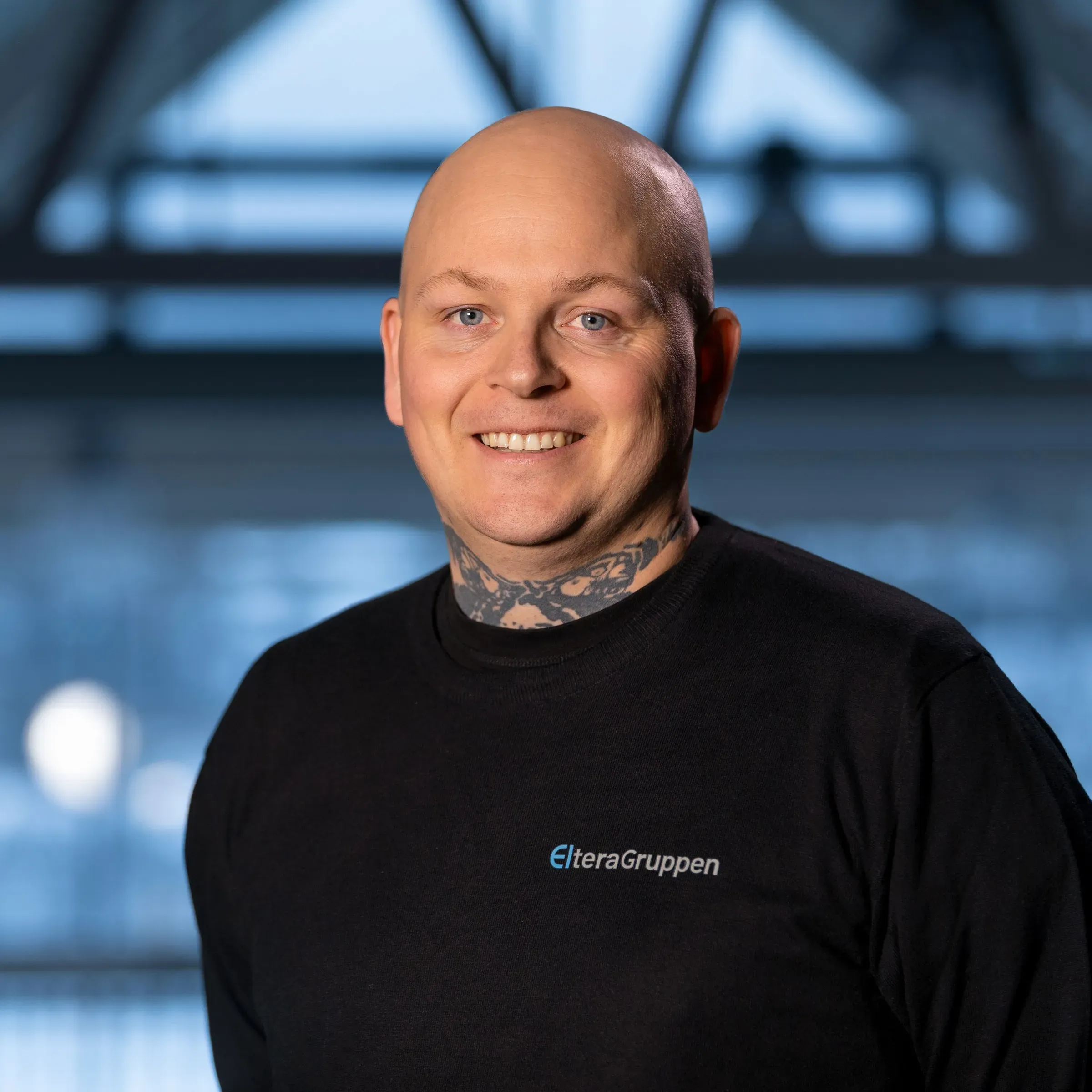 En smilende mann med barbert hode og synlig hals-tatovering, iført svart genser med logoen til Eltera