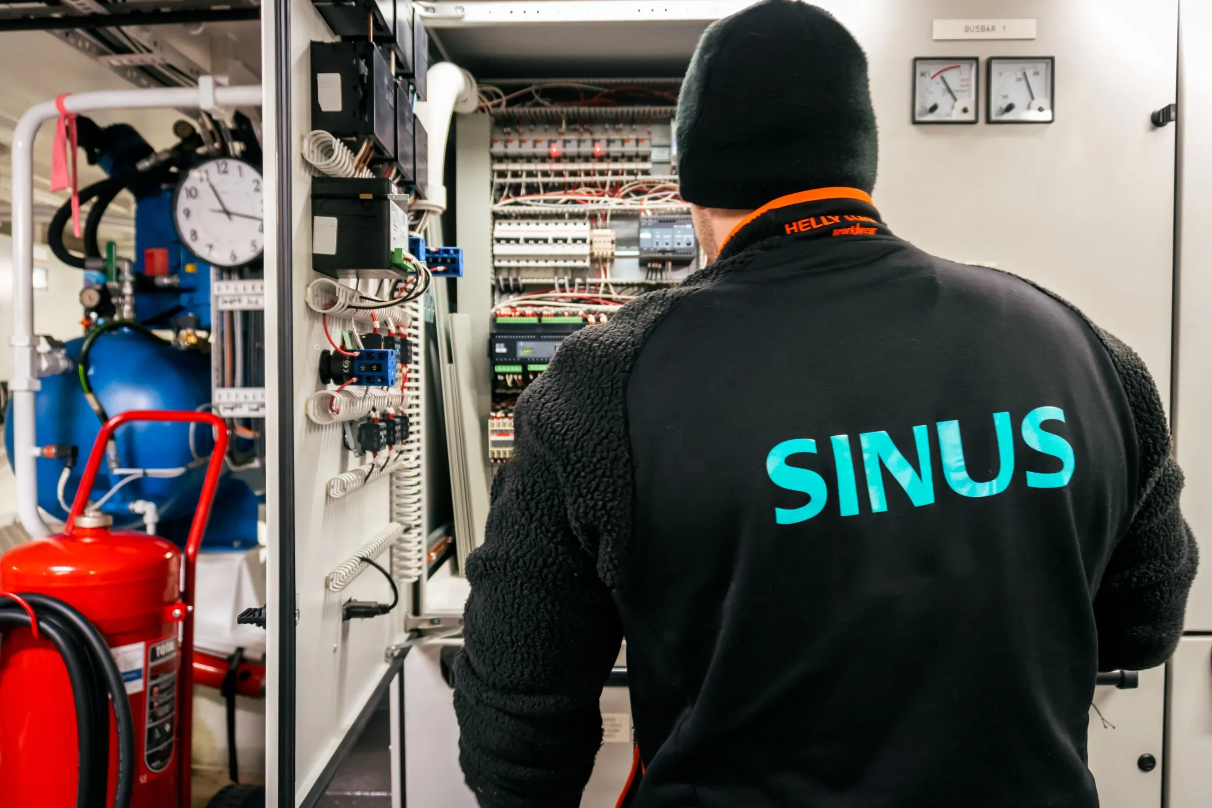 En person med en sort jakke med teksten 'SINUS' på ryggen står foran et elektrisk skap og jobber med elektriske komponenter.
