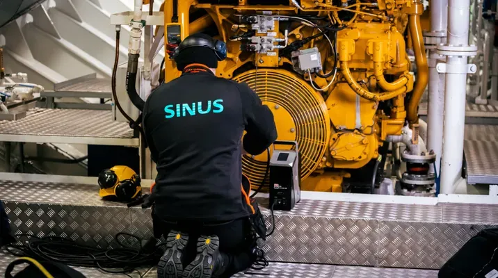 En tekniker iført svart arbeidsklær med teksten 'SINUS' jobber foran en stor, gul motormaskin ombord på et skip eller i et industrirom.