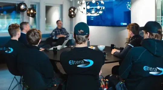 En gruppe personer sitter rundt et møtebord og ser på en presentasjon på en TV-skjerm. Flere har arbeidsklær med 'Eltera' logo på ryggen.