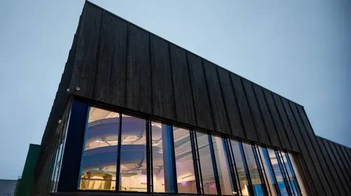 Ny svømmehall i mørk kledning med store glassfasader og lys innvendig, fotografert i skumringen.
