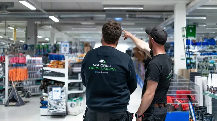 To personer i arbeidsklær står i en butikk full av verktøy og utstyr. Den ene peker noe ut for den andre.