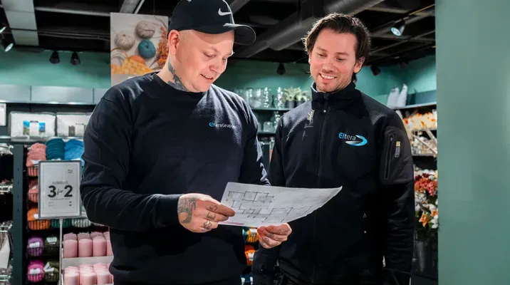 To menn i arbeidstøy står inne i en butikk og ser på et ark sammen, de smiler og diskuterer noe.