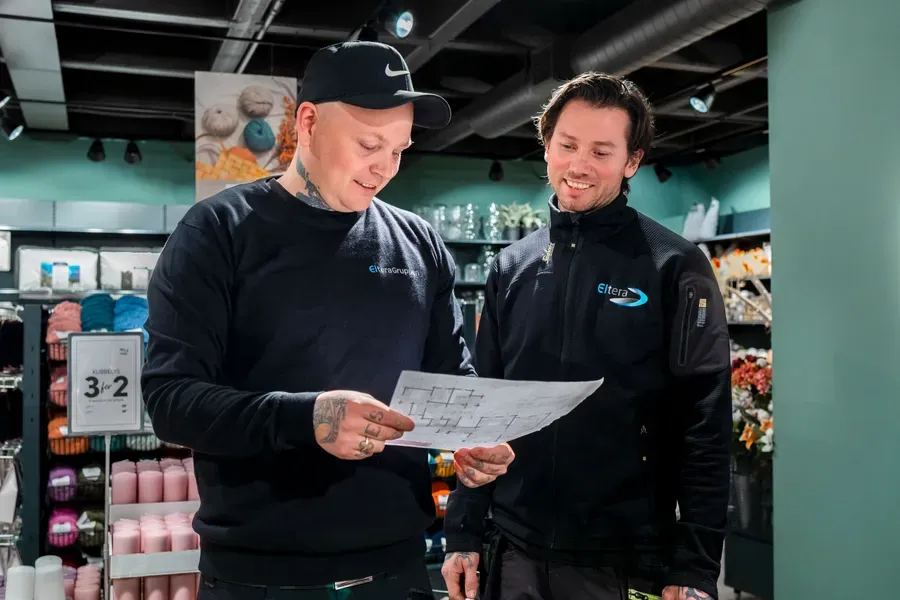 To menn i arbeidstøy står inne i en butikk og ser på et ark sammen, de smiler og diskuterer noe.