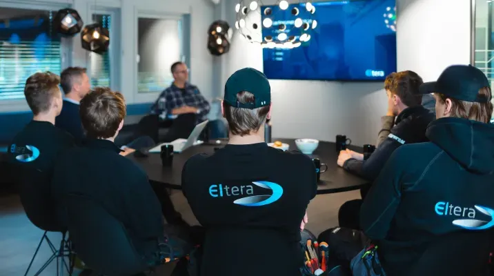 En gruppe personer sitter rundt et møtebord og ser på en presentasjon på en TV-skjerm. Flere har arbeidsklær med 'Eltera' logo på ryggen.