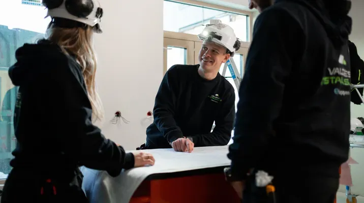 Tre personer i arbeidsklær og vernehjelmer ser på tegninger rundt et bord inne på en byggeplass.