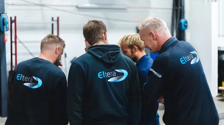 Fire menn med ryggen til står sammen i et industrielt innemiljø. De har sorte gensere eller jakker med logoen 'Eltera' på ryggen.