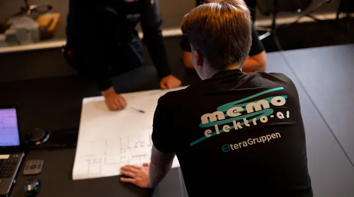 Tre personer diskuterer rundt et bord med tekniske tegninger. Personen nærmest kameraet har på seg en t-skjorte med logoen 'memo elektro as'.