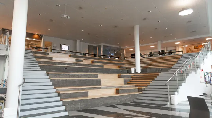 Et moderne innendørs amfiområde med brede trappeavsats og sitteplasser, omgitt av glassrekkverk og lyse søyler. Tepoer sitter gjerne folk til møter eller foredrag.