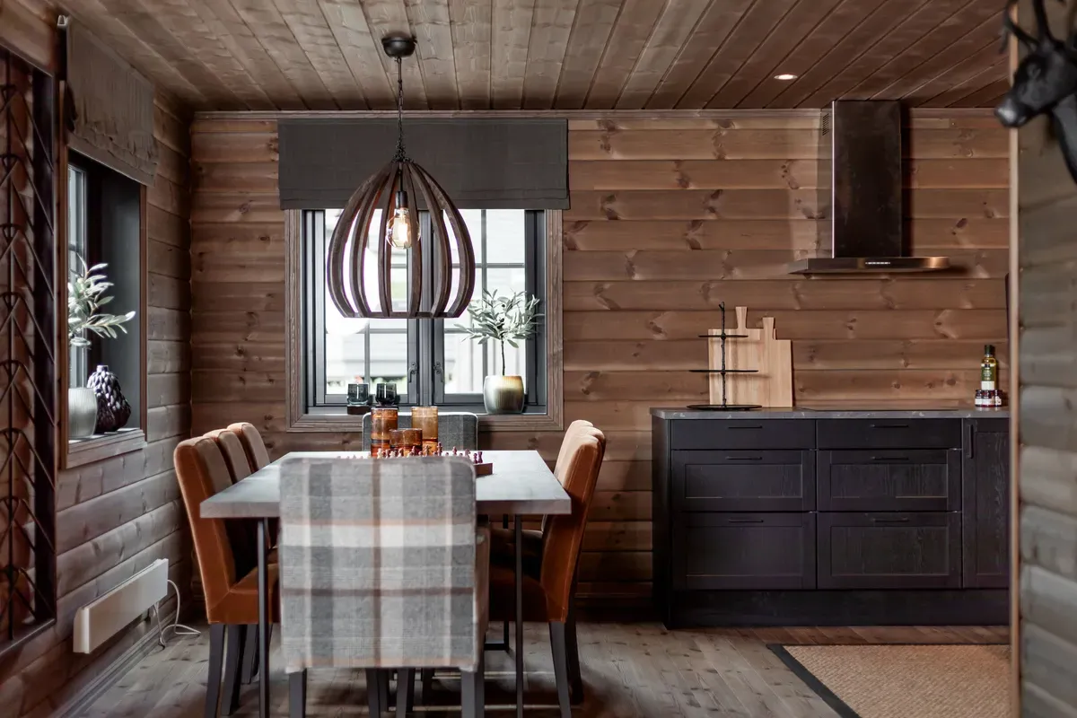 Et koselig kjøkken med tømmervegger, spisebord med beige stoler, mørke kjøkkenskap og moderne taklampe.