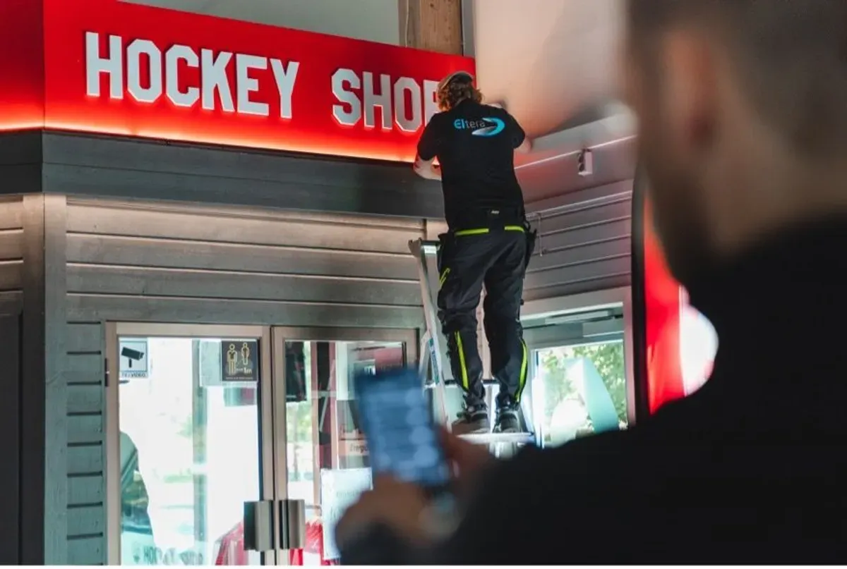 En person står på en stige og jobber med et skilt som sier 'Hockey Shop', mens en annen person holder en mobiltelefon i forgrunnen.