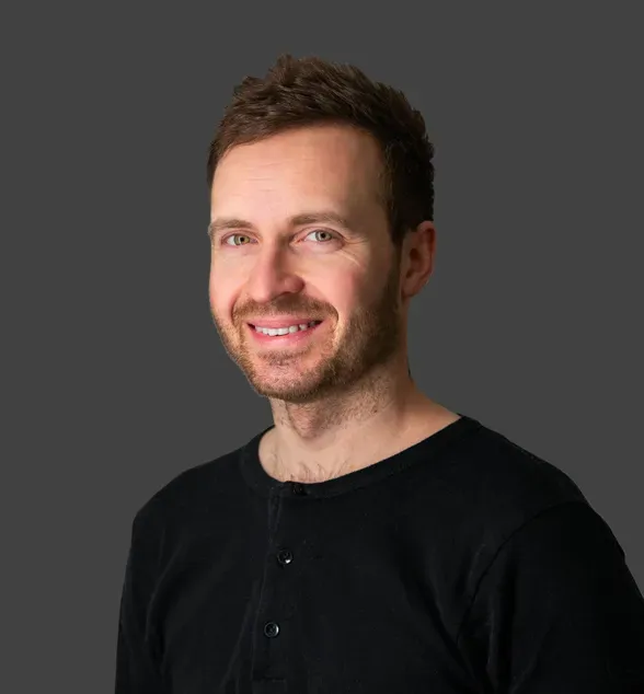Portrettbilde av en smilende mann med kort, brunt hår og skjegg, iført en svart genser, mot en ensfarget grå bakgrunn.