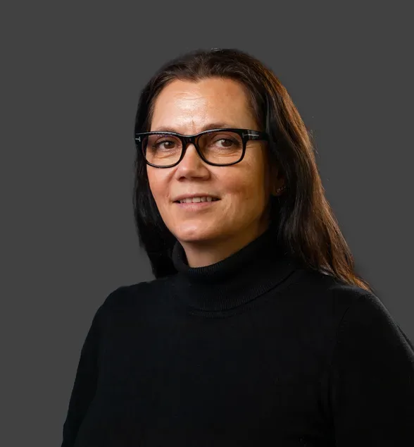 Kvinne med langt mørkt hår og briller, iført svart genser. Hun står foran en nøytral, mørk bakgrunn.