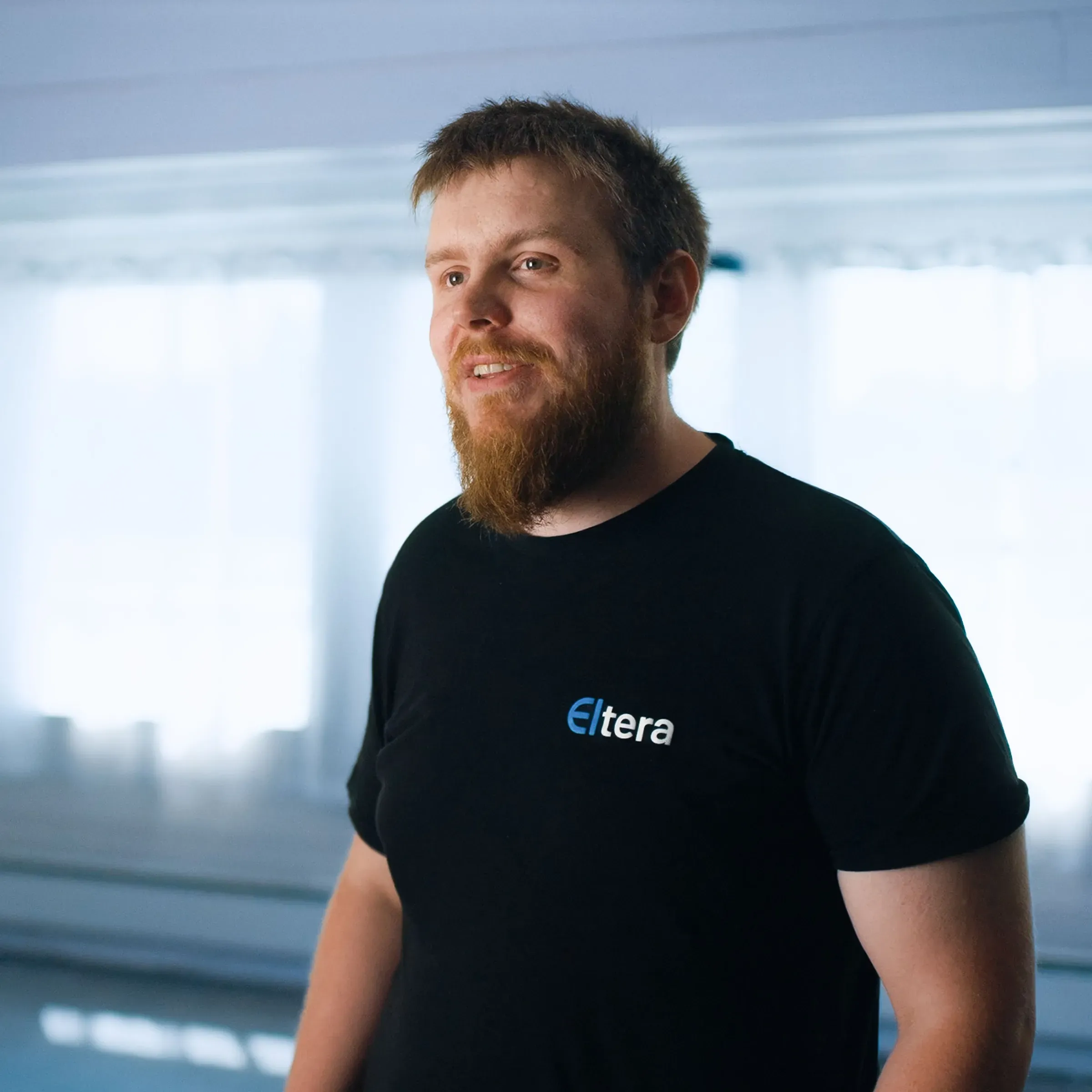 En mann med skjegg står innendørs, iført en svart t-skjorte med Eltera-logoen.