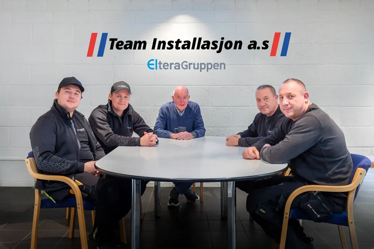 Fem menn sitter rundt et bord foran en vegg med logoen 'Team Installasjon a.s' og 'ElteraGruppen'.