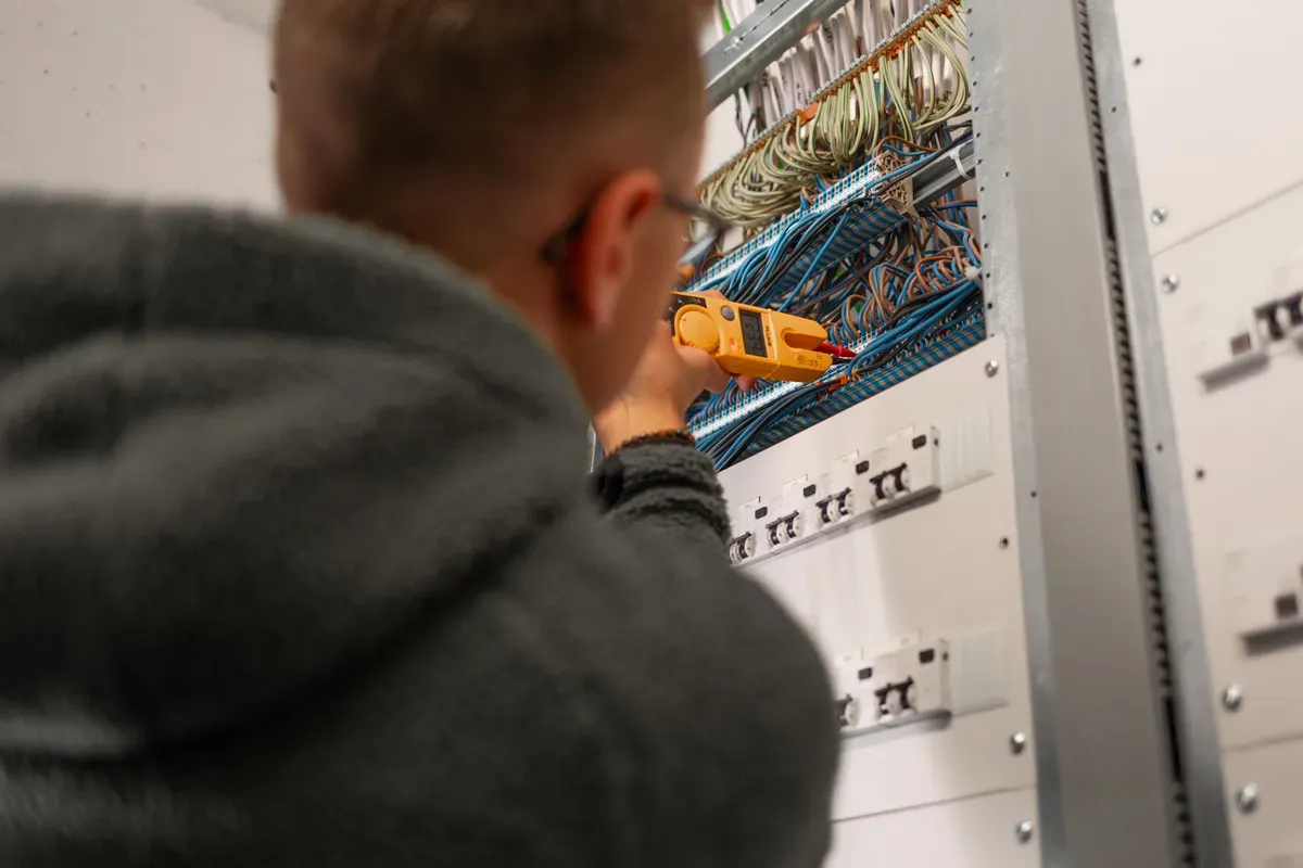 En person som arbeider med elektrisk utstyr og ledninger i et sikringsskap, sett bakfra.