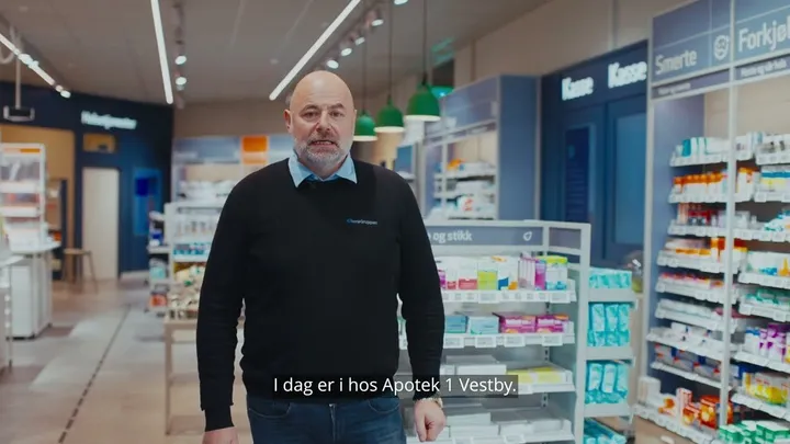 En mann med svart genser og jeans står i midten av et apotek, omgitt av hyller med medisiner og produkter. Han ser rett mot kameraet. Norsk tekst nederst sier: 'I dag er I hos Apotek 1 Vestby.'