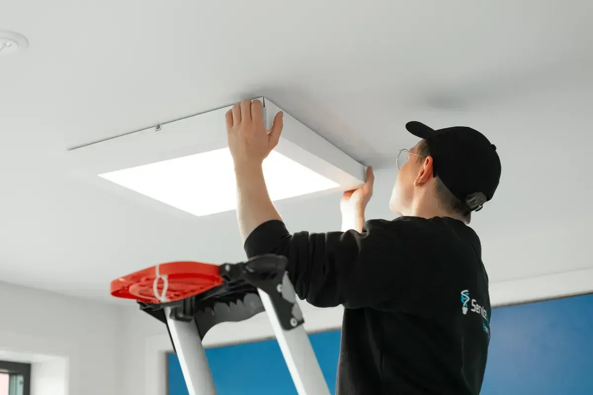 En person på en stige installerer en firkantet taklampe i et rom med hvitt tak og blå vegg.