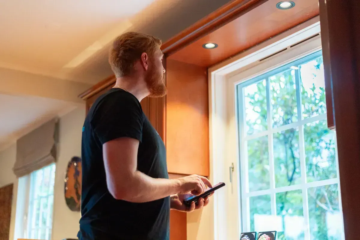 En mann med skjegg står inne og ser ut av et vindu mens han holder en mobiltelefon.