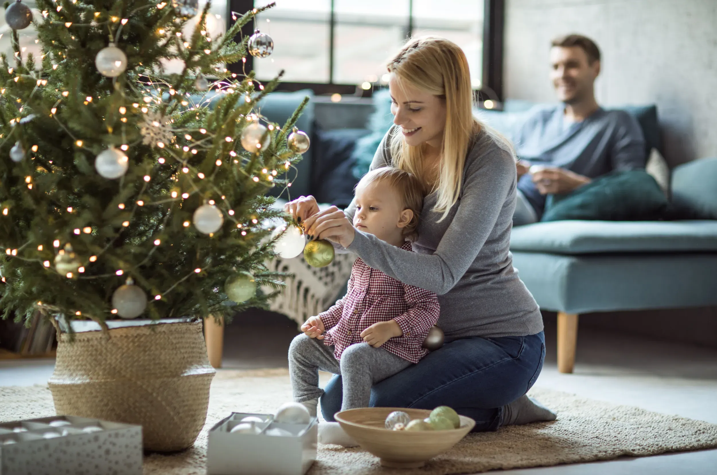 mor og barn som pynter juletre og en mann som sitter i sofaen