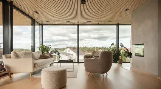 Lyst og moderne stue med store vinduer, lyse tremøbler og utsikt mot naturen utenfor.