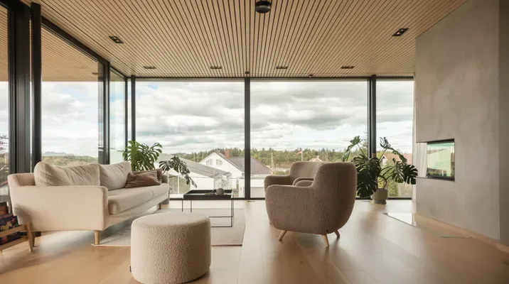 Lyst og moderne stue med store vinduer, lyse tremøbler og utsikt mot naturen utenfor.