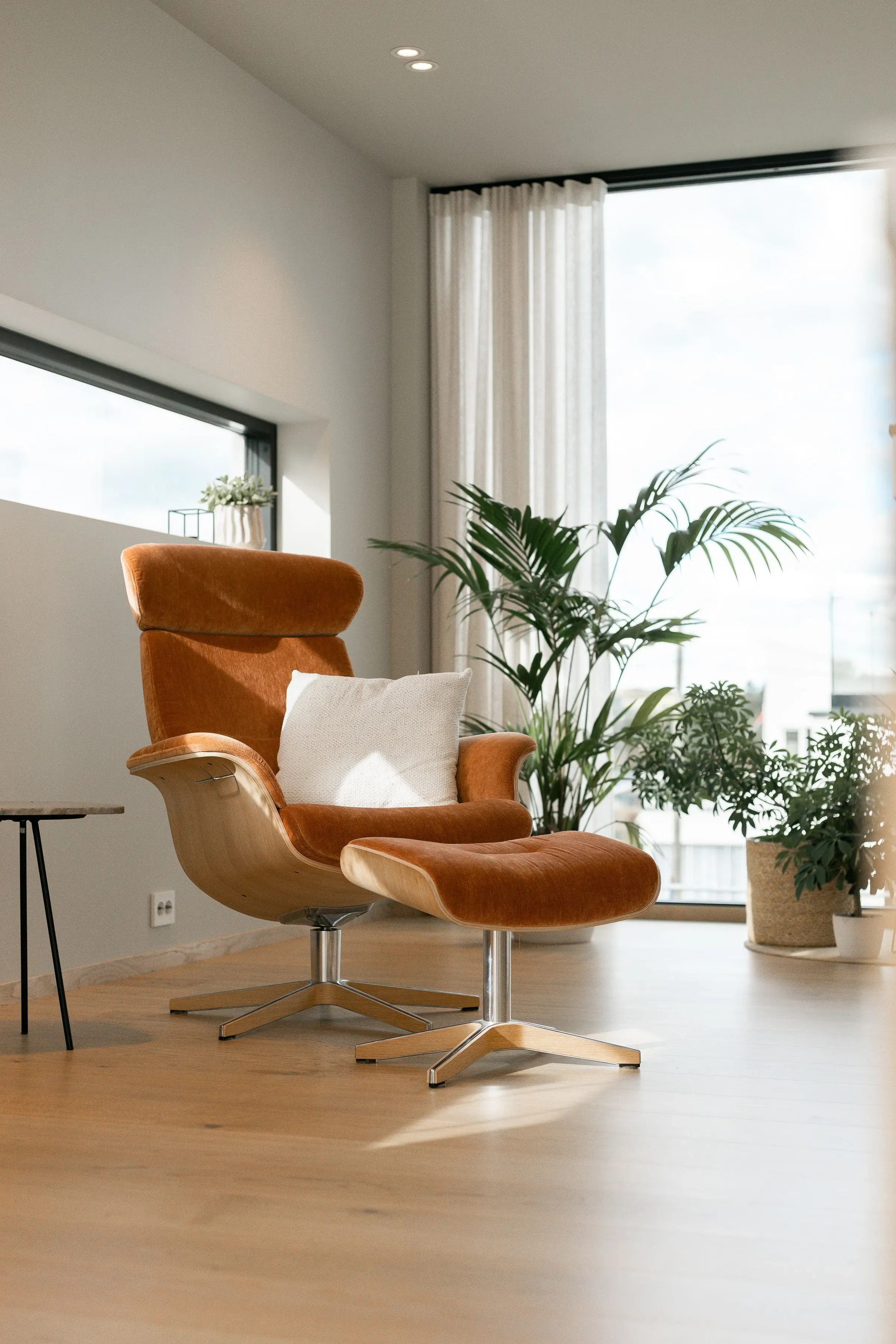 En moderne stue med en oransje lenestol og en fotskammel, en hvit pute, grønne planter i bakgrunnen og store vinduer med gardiner.