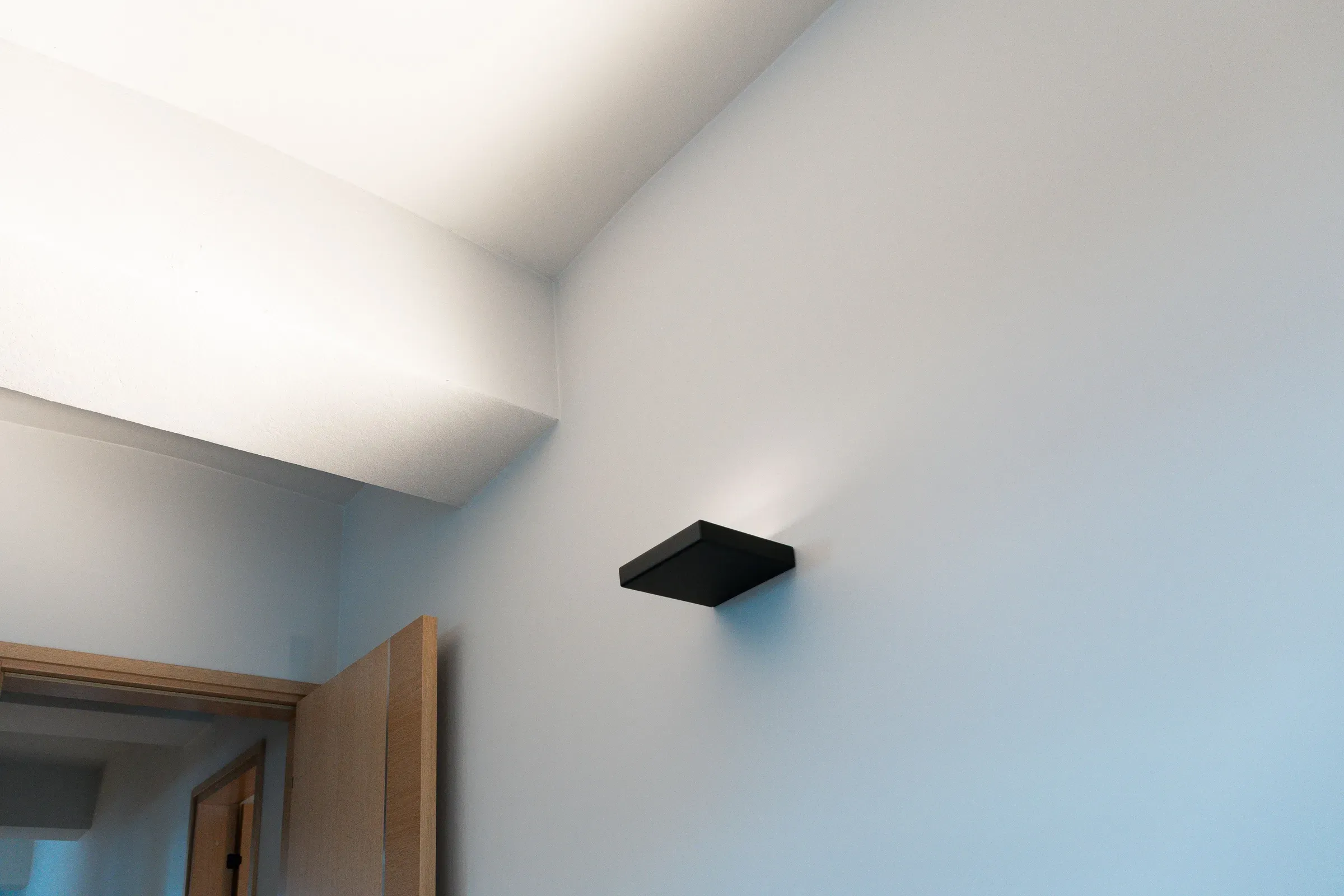 En moderne vegglampe med svart rektangulært design montert på en blå vegg, delvis åpnet dør til venstre i bildet.